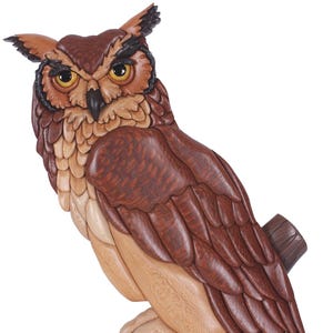Op de afbeelding: Houten uil sculptuur op een tak. De uil is gedetailleerd gesneden in bruin, beige en zwart. De gele ogen en klauwen zijn zichtbaar. Een decoratief object.