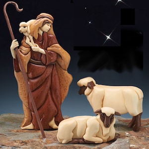 May include: A wooden figurine of a shepherd carrying a lamb, with two other lambs lying on a stone surface. The shepherd is wearing a brown robe and a brown hat. The lambs are white with brown heads.
