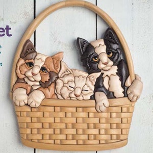 Op de afbeelding: Drie houten gesneden kittens in een mand. De kittens zijn een calico, een wit kitten met bruine ogen en een zwart-wit kitten. De mand is gemaakt van gevlochten hout en heeft een bruine vlek.
