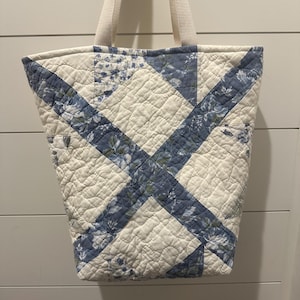 May include: Quilted tote bag with a cream background and blue floral accents. The bag features a quilted design with a cream-colored handle. The bag's design includes a blue floral pattern and a cross-stitch pattern.