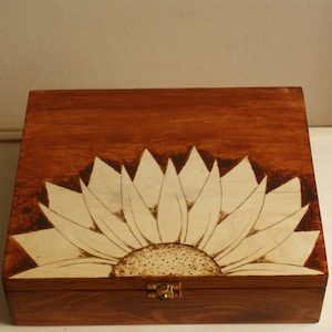 May include: A wooden box with a sunflower design. The box is stained a dark brown color, with a lighter wood sunflower design burned into the top. The sunflower has a detailed center and pointed petals. A small metal clasp secures the box.