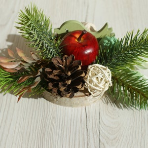 Może przedstawiać: Rúsztyczny świąteczny element dekoracyjny z czerwonym jabłkiem, szyszkami i zielonymi gałązkami na drewnianym plastrze.