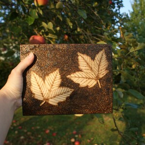 May include: A wooden plaque with two burnt-in images of maple leaves. The plaque is dark brown and has a rough texture.