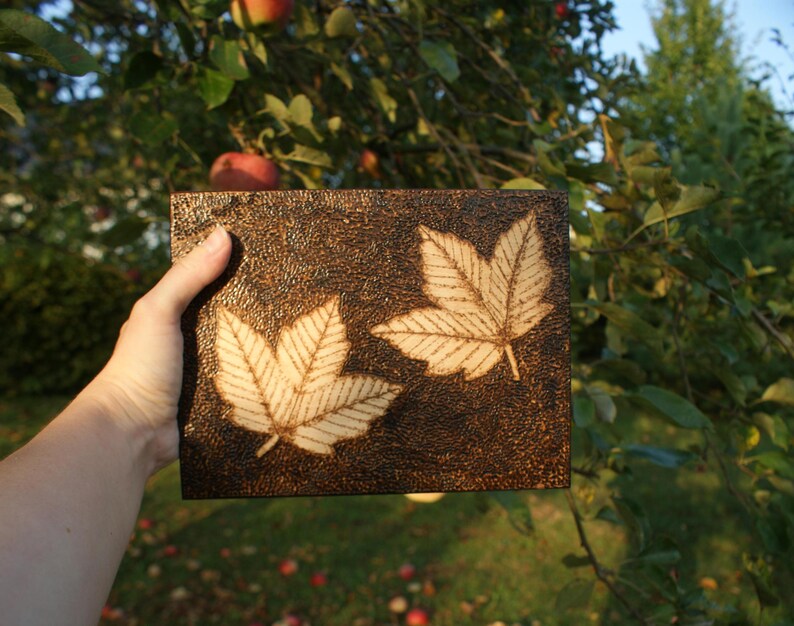 May include: A wooden plaque with two burnt-in images of maple leaves. The plaque is dark brown and has a rough texture.