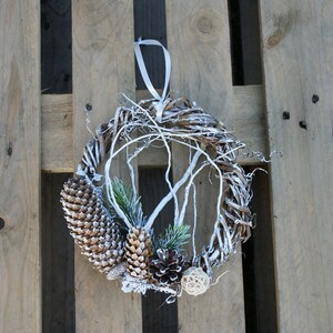 Puede incluir: Una corona blanca de invierno con ramas de pino, pi&ntilde;as y una cinta blanca. La corona est&aacute; colgada en una paleta de madera.
