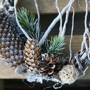 Puede incluir: Una corona de ramas blanqueadas decorada con ramas de pino, pi&ntilde;as y una bola de mimbre natural.