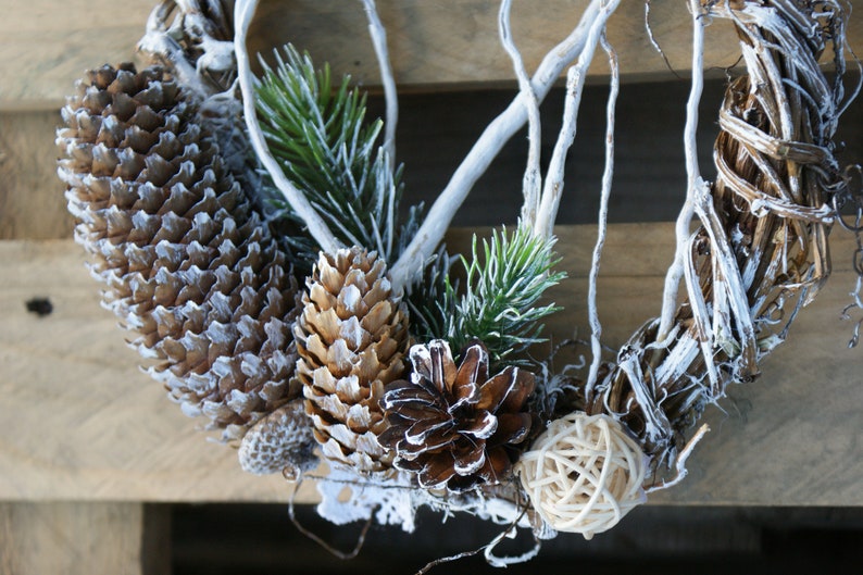 Puede incluir: Una corona de ramas blanqueadas decorada con ramas de pino, pi&ntilde;as y una bola de mimbre natural.