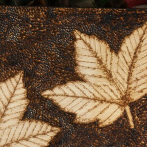 May include: A close-up of a wooden plaque with two stylized leaf designs burned into the surface. The leaves are light brown and the background is dark brown.