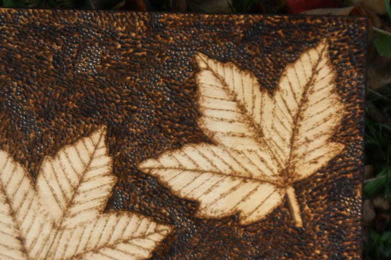 May include: A close-up of a wooden plaque with two stylized leaf designs burned into the surface. The leaves are light brown and the background is dark brown.
