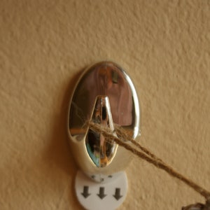 May include: A silver hook with a string attached to it. The hook is mounted on a wall with a white sticker with three arrows pointing down.