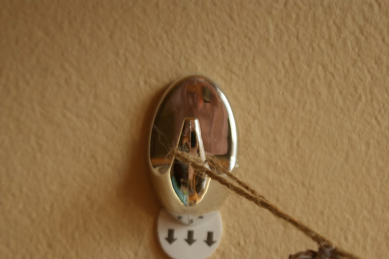 May include: A silver hook with a string attached to it. The hook is mounted on a wall with a white sticker with three arrows pointing down.