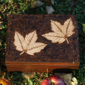 May include: A wooden box with a dark brown finish and a woodgrain pattern. The box has a hinged lid and is decorated with two white leaf designs burned into the surface.