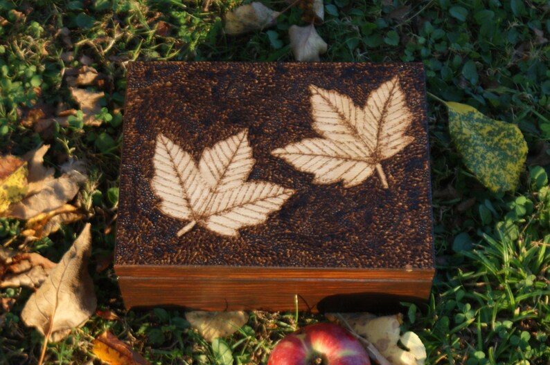 May include: A wooden box with a dark brown finish and a woodgrain pattern. The box has a hinged lid and is decorated with two white leaf designs burned into the surface.