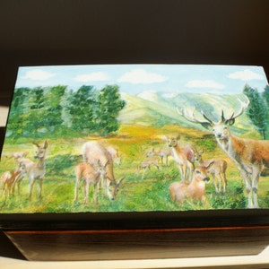Puede incluir: Una caja de madera con una escena pintada de un bosque con ciervos. La caja tiene un marco de madera marrón oscuro y una base de madera marrón claro. La escena pintada presenta un grupo de ciervos en un campo de hierba con árboles y montañas en el fondo.