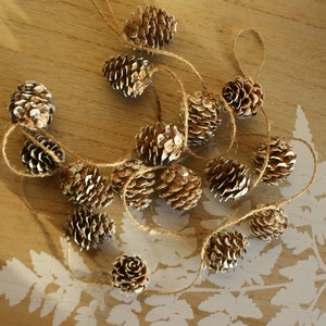 May include: A string of gold-painted pine cones strung together with twine. The pine cones are arranged in a circular pattern on a wooden surface.