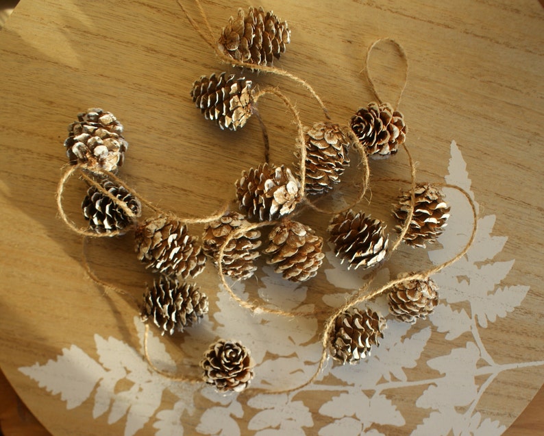 May include: A string of gold-painted pine cones strung together with twine. The pine cones are arranged in a circular pattern on a wooden surface.