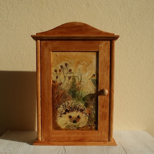 Puede incluir: Un armario de madera con una puerta con bisagras y un pomo. La puerta presenta una imagen pintada de un erizo en un entorno de bosque.