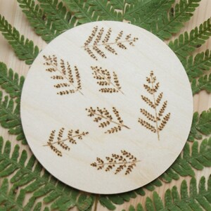 May include: A round wooden coaster with a design of seven fern leaves burned into the surface. The coaster is surrounded by green fern leaves.