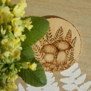 May include: A wooden coaster with a burnt design of two mushrooms and ferns. The coaster is round and has a light brown color.