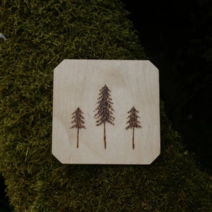 Può includere: Un sottobicchiere di legno con un design di tre pini bruciati sulla superficie.