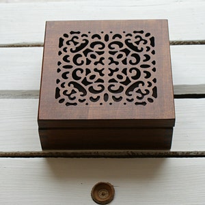 May include: A dark brown wooden box with a decorative cutout design on the lid. The box is sitting on a white surface.