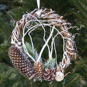 Puede incluir: Una corona de ramas blanqueadas con ramas de pino y pi&ntilde;as, decorada con una rama blanqueada y una peque&ntilde;a bola de color natural.