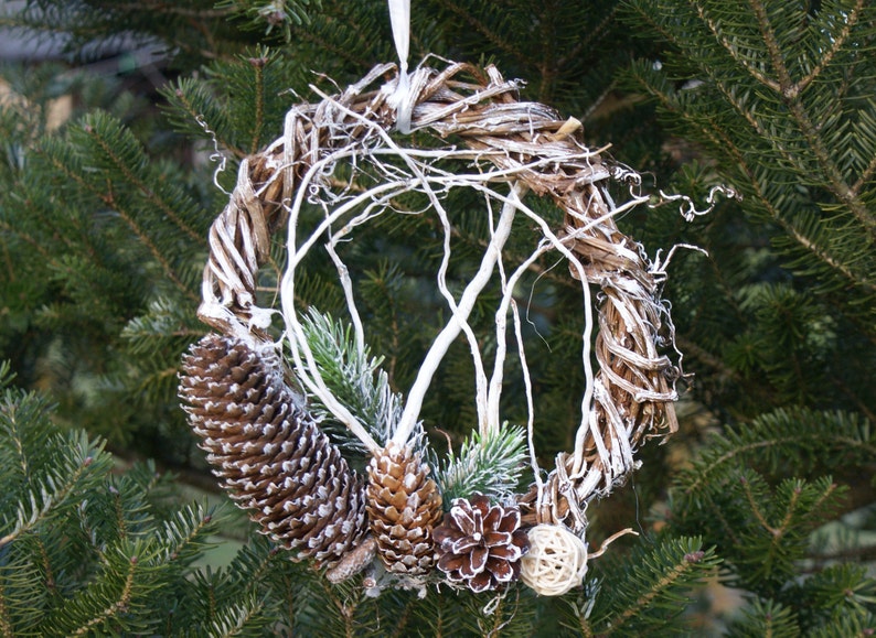 Puede incluir: Una corona de ramas blanqueadas con ramas de pino y pi&ntilde;as, decorada con una rama blanqueada y una peque&ntilde;a bola de color natural.
