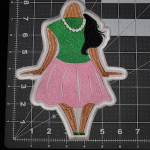 May include: An embroidered iron-on patch depicting a cartoon woman wearing a green shirt, pink skirt, and a white pearl necklace. The woman has brown skin, black hair, and is standing with her arms outstretched.