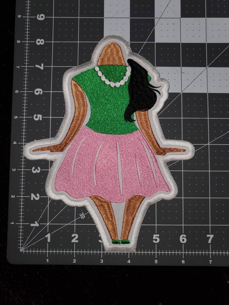 May include: An embroidered iron-on patch depicting a cartoon woman wearing a green shirt, pink skirt, and a white pearl necklace. The woman has brown skin, black hair, and is standing with her arms outstretched.