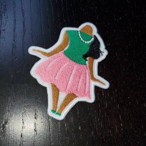 May include: An embroidered patch depicting a stylised figure of a woman wearing a pink skirt, green top, and brown skin. The figure is standing with her arms outstretched.