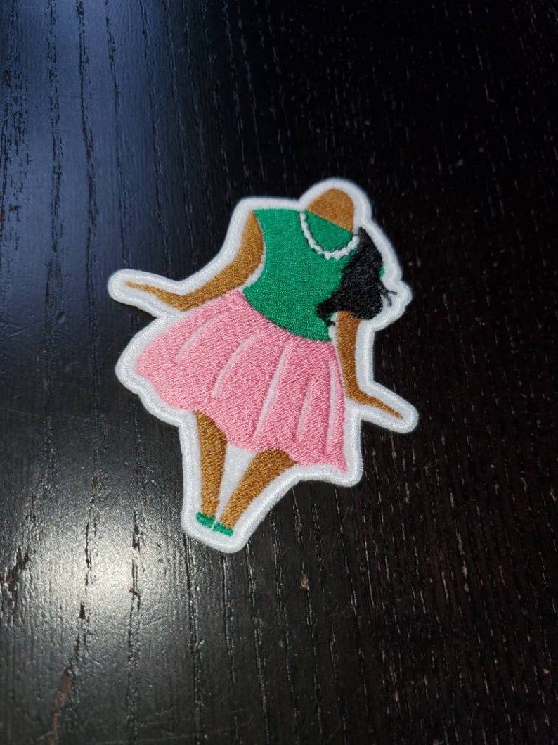 May include: An embroidered patch depicting a stylised figure of a woman wearing a pink skirt, green top, and brown skin. The figure is standing with her arms outstretched.