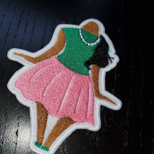 May include: An embroidered patch depicting a woman wearing a pink skirt and green top. The woman has brown skin and black hair. The patch is outlined in white.