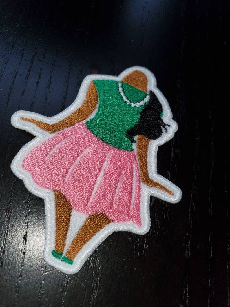 May include: An embroidered patch depicting a woman wearing a pink skirt and green top. The woman has brown skin and black hair. The patch is outlined in white.