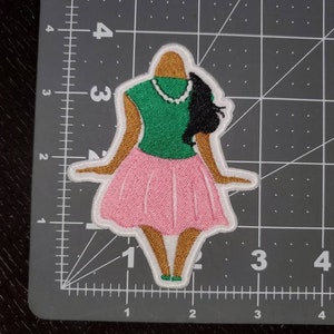 May include: Embroidered patch of a woman wearing a green shirt, pink skirt, and a pearl necklace. Her hair is black and pulled back in a ponytail.