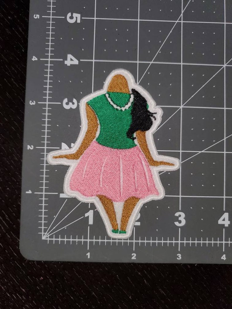 May include: Embroidered patch of a woman wearing a green shirt, pink skirt, and a pearl necklace. Her hair is black and pulled back in a ponytail.