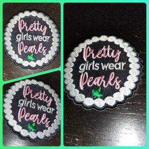May include: Embroidered patch with the text "Pretty girls wear Pearls" in pink script. The patch has a navy blue background, a white pearl-like border, and a small green leaf design. The patch is round with a scalloped edge.