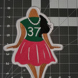 May include: Embroidered patch featuring a cartoon woman wearing a green shirt with the number 37, a pink skirt, and a pearl necklace. The patch has a white outline.