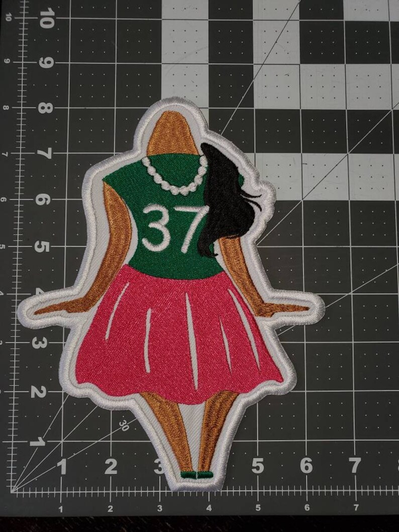 May include: Embroidered patch featuring a cartoon woman wearing a green shirt with the number 37, a pink skirt, and a pearl necklace. The patch has a white outline.