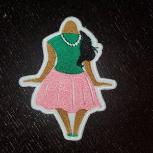 May include: An embroidered patch depicting a woman wearing a green top, pink skirt, and a white pearl necklace. The woman has brown skin and black hair.