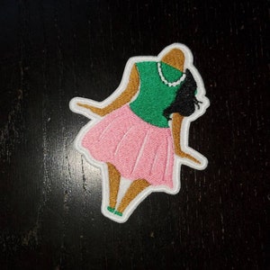 May include: Embroidered patch of a woman wearing a green top and pink skirt. The woman has brown skin and black hair.