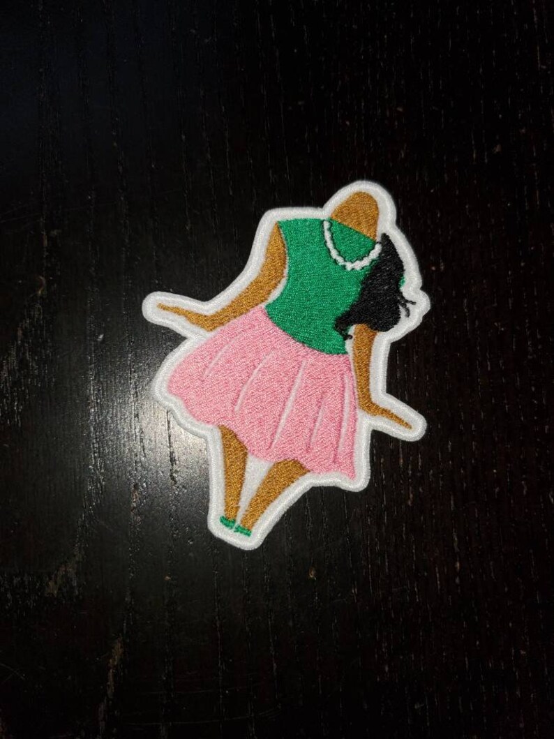 May include: Embroidered patch of a woman wearing a green top and pink skirt. The woman has brown skin and black hair.