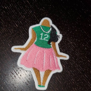 May include: An embroidered patch depicting a cartoon woman wearing a green shirt with the number "12" on it, a pink skirt, and a pearl necklace.