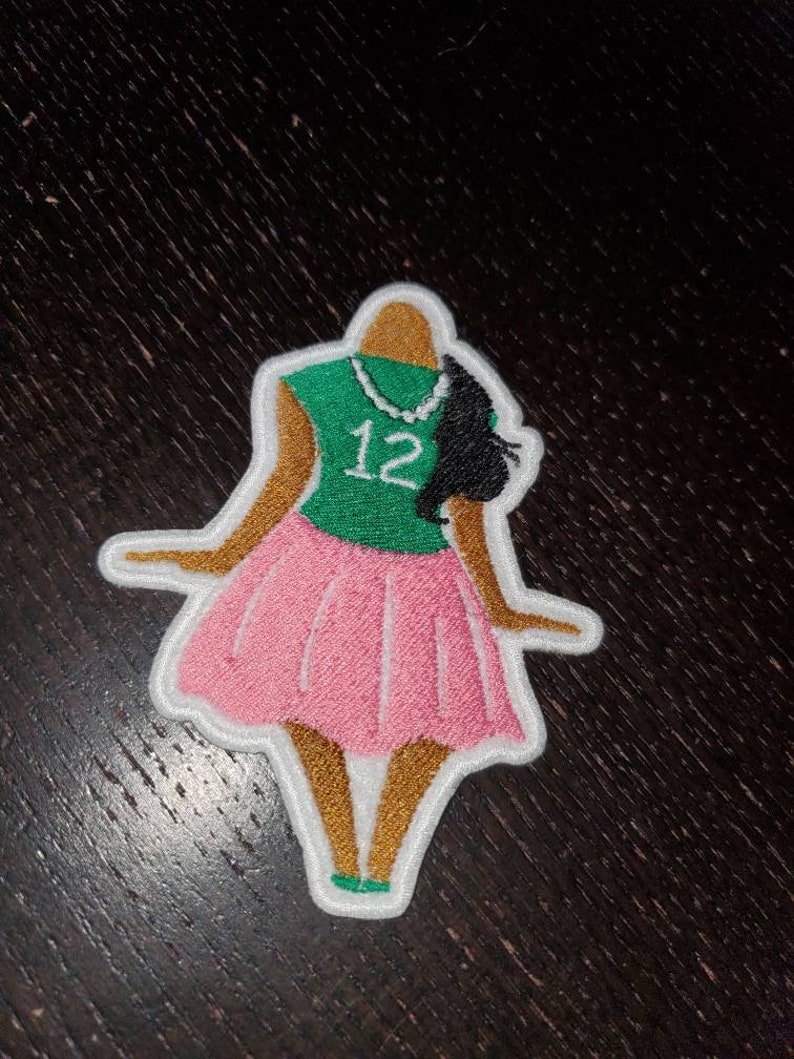 May include: An embroidered patch depicting a cartoon woman wearing a green shirt with the number "12" on it, a pink skirt, and a pearl necklace.