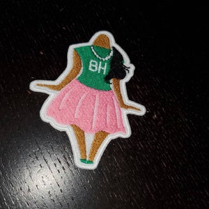 May include: An embroidered patch depicting a cartoon figure of a woman wearing a green shirt with the letters "BH" on it, a pink skirt, and a pearl necklace. The figure has brown skin and black hair.