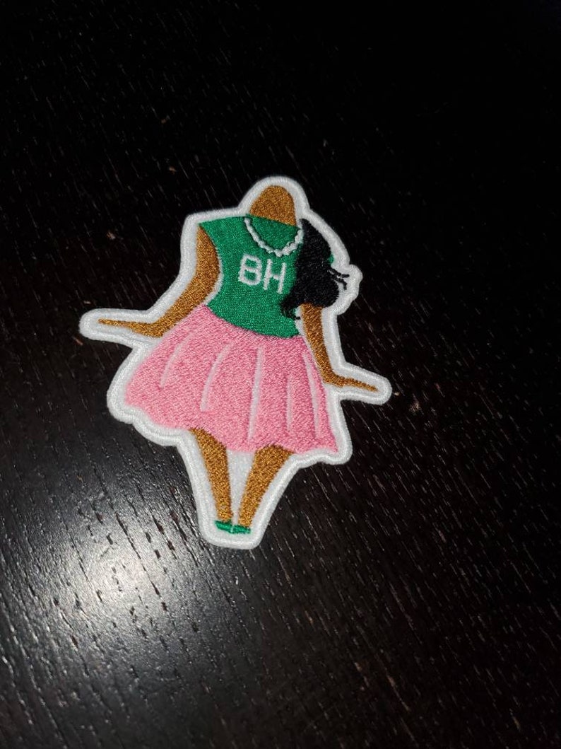 May include: An embroidered patch depicting a cartoon figure of a woman wearing a green shirt with the letters "BH" on it, a pink skirt, and a pearl necklace. The figure has brown skin and black hair.