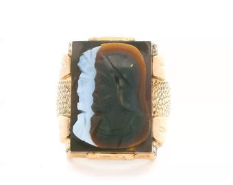 May include: A gold ring featuring a rectangular cameo with a dark brown background and two carved profiles in light blue and brown. The band has a textured, cross-hatched pattern on the sides, and the setting is polished gold metal.