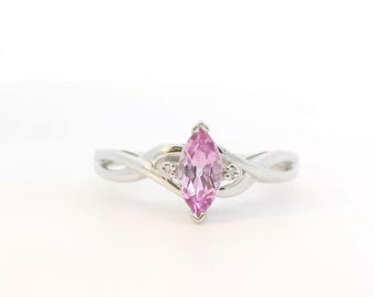 May include: A silver ring featuring a marquise-cut pink gemstone at the center, flanked by two small clear stones on a polished band with a twisted, crossover design that tapers toward the back of the ring.
