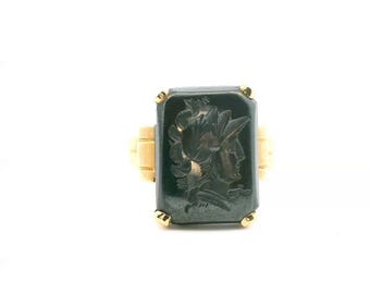 May include: A gold ring featuring a rectangular, dark gray hematite stone carved with a profile of a helmeted figure. The stone is held in place by four small gold prongs at the corners, set upon a simple, polished gold band with side detailing.