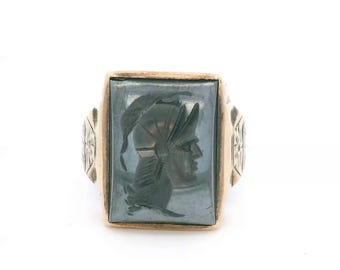May include: A gold ring featuring a rectangular, dark gray hematite intaglio stone. The stone is carved with the profile of a warrior wearing a crested helmet. The gold band displays intricate engraved detailing on both sides of the setting.