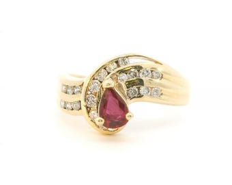 May include: A gold ring featuring a central pear-shaped red gemstone surrounded by a cluster of small round clear stones. The band has a bypass design with two rows of channel-set clear stones extending from the center setting.